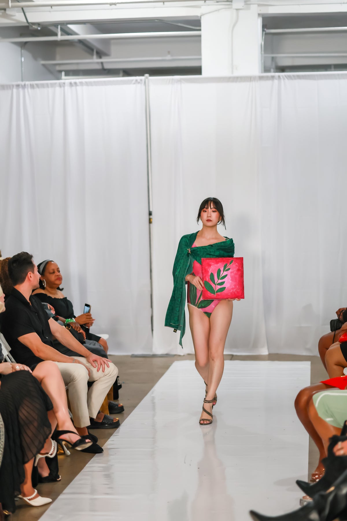 Full-length view of a model in a pink one piece swimsuit with a green scarf draped over her shoulders on a white runway